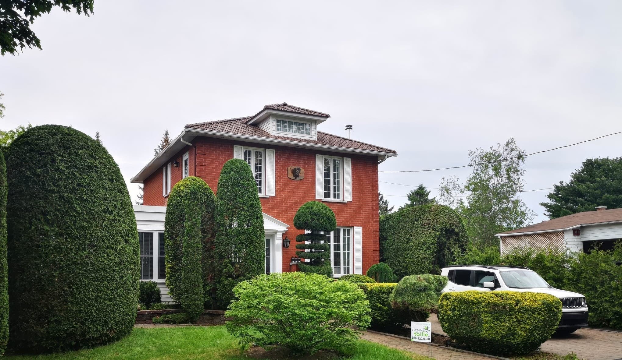 Devant d'une maison de brique rouge entièrement sculpté : haie massive arrondie, cèdres colonnaires, pin taillé en étages et boules de buis